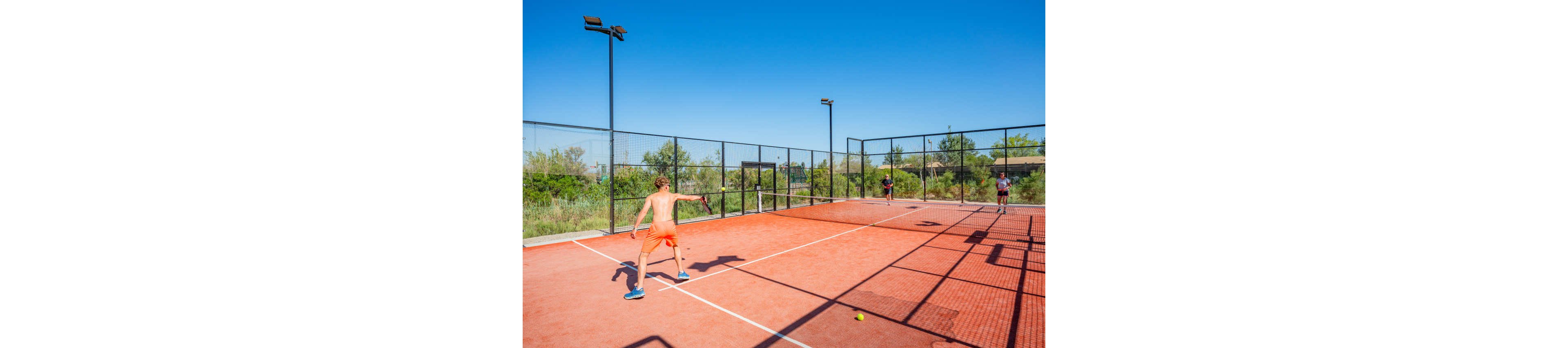 Several people play padel on an outdoor red court under a clear blue sky. The fenced court is surrounded by greenery and tall light poles. One player in the foreground prepares to hit the ball while others wait near the opposite side. Strong sunlight creates sharp shadows on the court, emphasizing the bright, summery atmosphere.