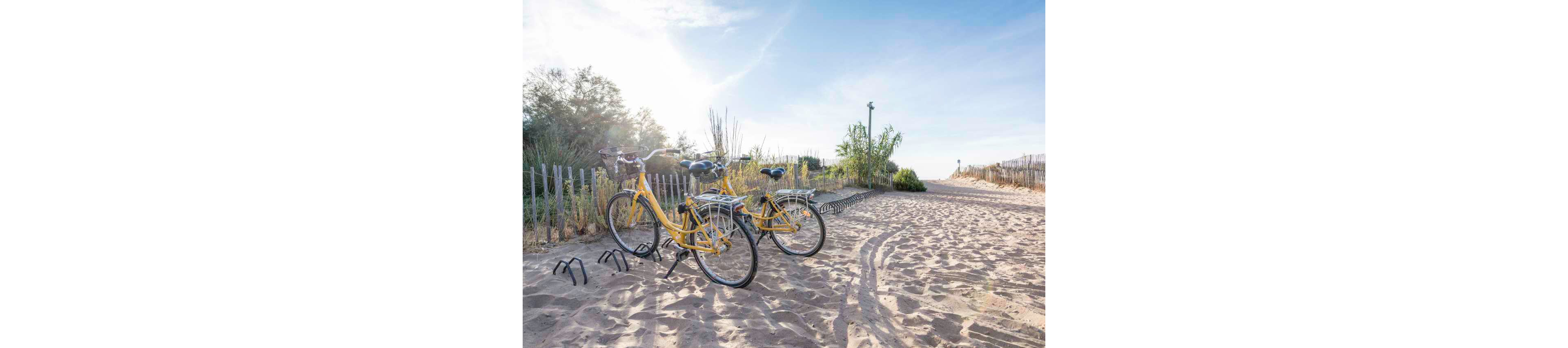 Two yellow bicycles are parked on a sandy path leading toward the beach. Wooden fences and sparse vegetation line the pathway, suggesting a coastal dune environment. The sky is clear and bright, creating a relaxed, summery atmosphere. Tire tracks and long shadows in the sand emphasize the warm, late-afternoon light.