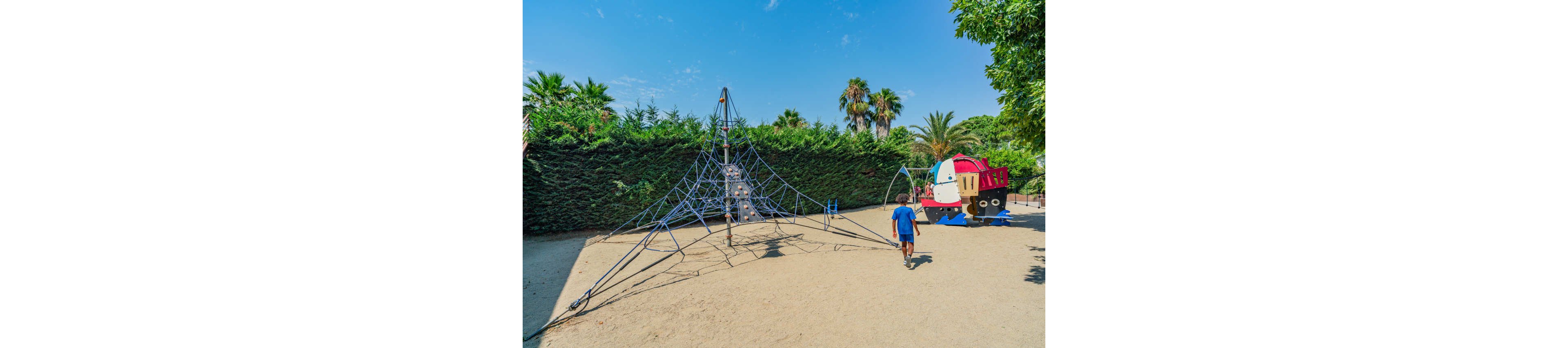 A young boy walks toward a large rope climbing structure in a sunny outdoor playground. The play area features a tall web-like frame with a central tower set on sandy ground. In the background, a colorful train-shaped play structure adds a playful focal point. Lush green hedges and palm trees surround the playground under a clear blue sky.
