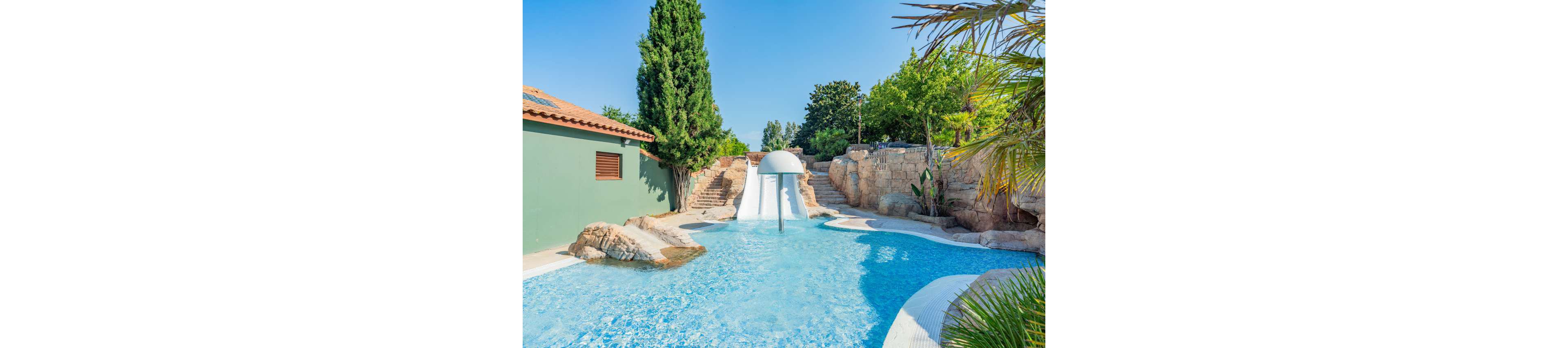 An outdoor swimming pool features a white curved waterslide ending in clear blue water. Stone steps and rock formations surround the pool, creating a natural, resort-like atmosphere. Lush green trees and tropical plants frame the scene under a bright blue sky. A nearby building with a terracotta roof adds to the warm, Mediterranean feel of the setting.