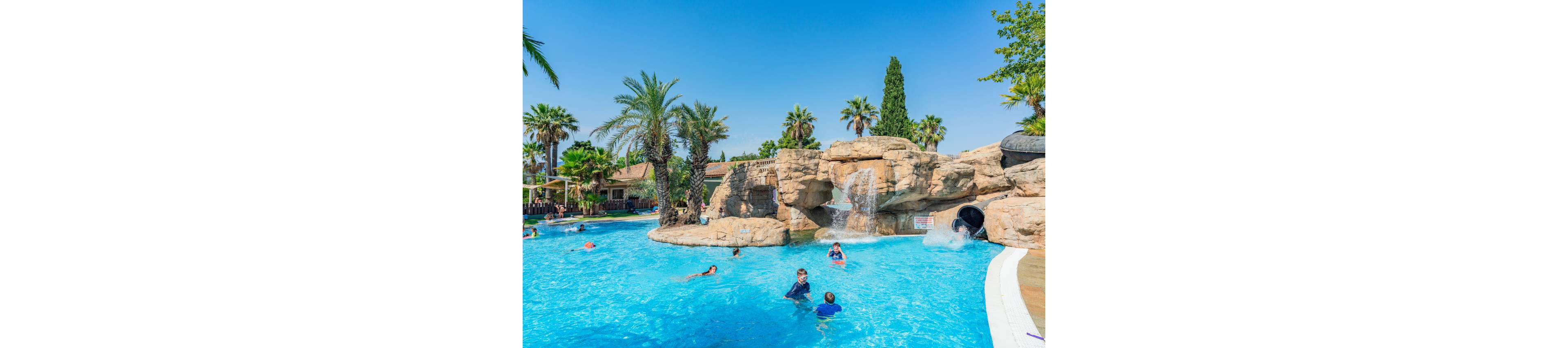Children and adults swim and play in a bright blue resort pool. The pool features artificial rock formations with waterfalls and a waterslide. Tall palm trees and clear blue sky create a tropical vacation atmosphere. People relax and enjoy the sunny weather around the water attraction.