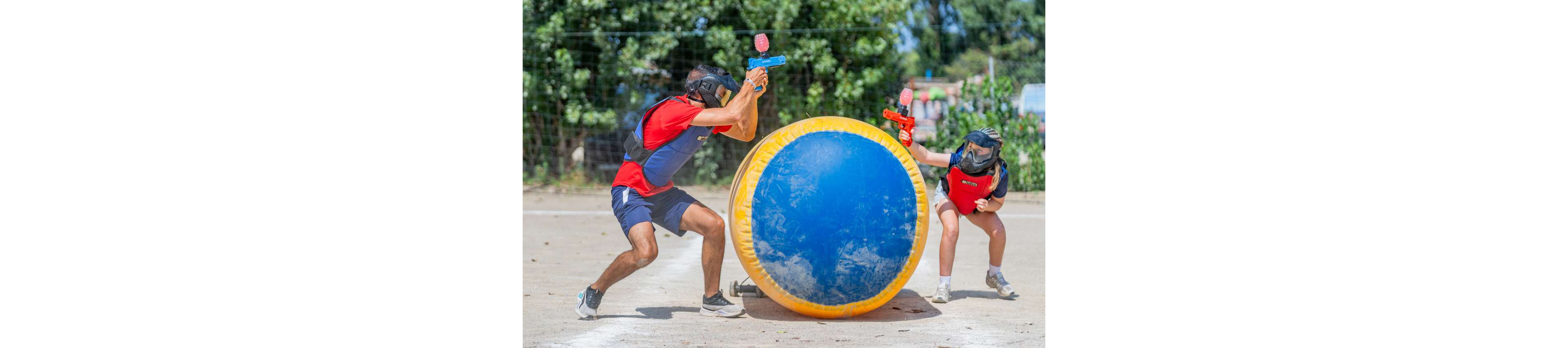 Two players wearing protective vests and masks take cover behind a large round inflatable bunker during an outdoor game. One player stands on the left aiming a blue toy gun, while the other crouches on the right with a red toy gun. The scene takes place on a sunny day on a dirt field with green trees and a fence in the background. Bright colors and dynamic poses emphasize action, teamwork, and recreation.