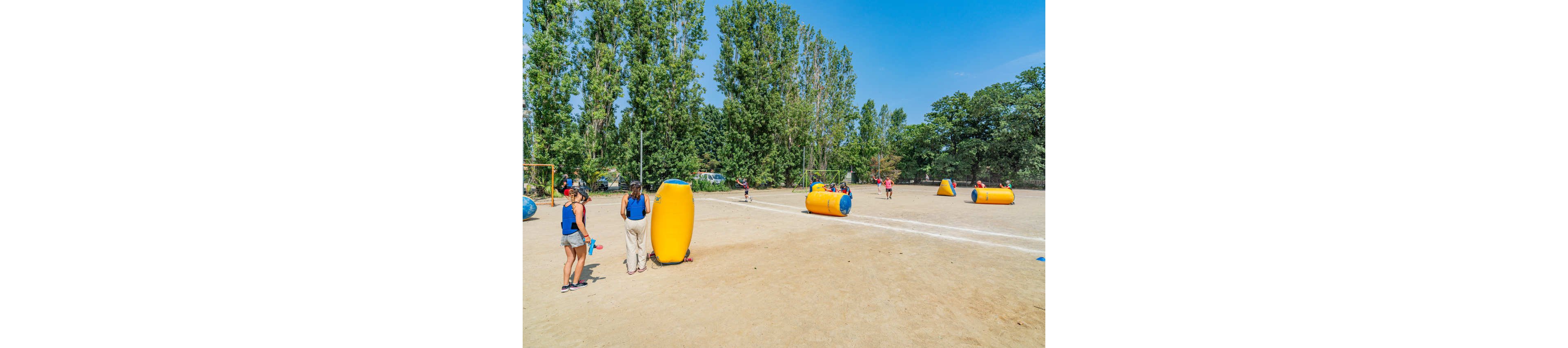 A group of people play an outdoor game on a large sandy field using bright yellow inflatable obstacles. Some participants stand on the sidelines while others run and interact around the inflatables in the center of the field. Tall green trees border the area under a clear blue sky, suggesting a warm sunny day. The scene conveys an active, recreational atmosphere suitable for team-building or leisure activities.