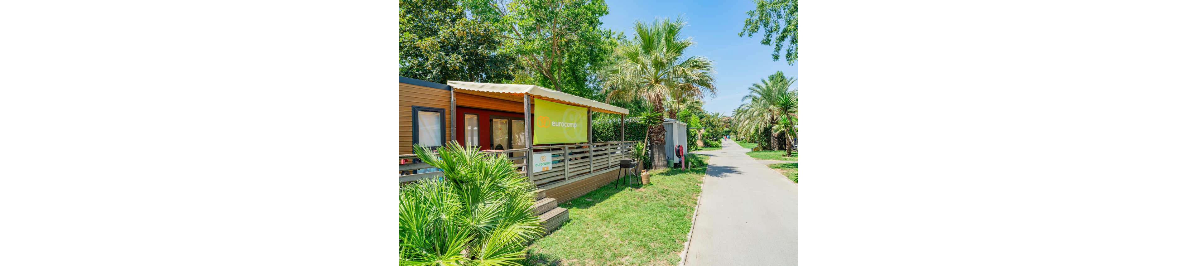 A modern mobile home with a covered wooden terrace stands beside a paved path in a lush campsite. The terrace features outdoor seating and is surrounded by green grass, palm trees, and other tropical plants. A yellow sign with the word "eurocamp" is clearly visible on the front of the terrace. In the background, a person walks along the path under the bright daylight sky. The overall scene feels relaxed, summery, and family friendly.