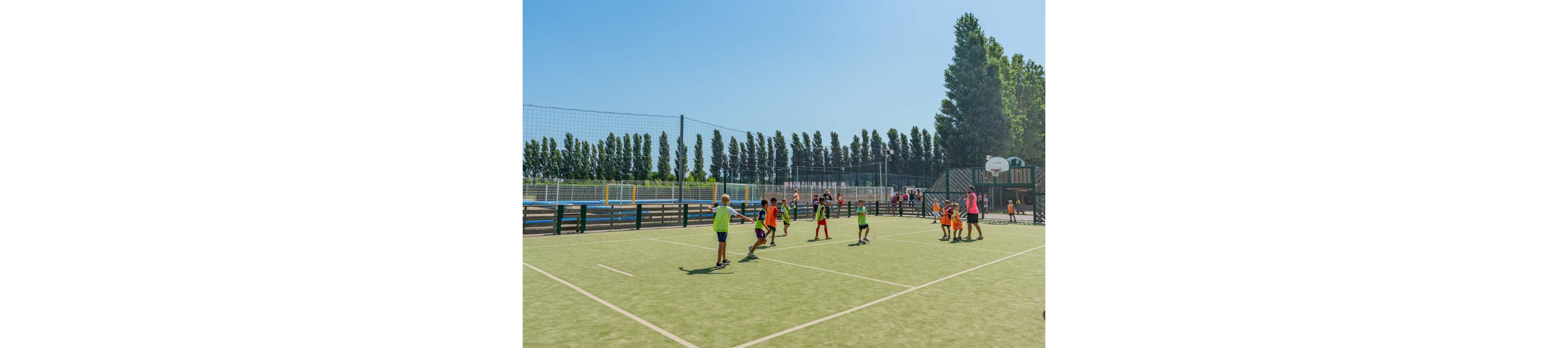 A group of children play a game on an outdoor sports court under a clear blue sky. The court is surrounded by tall trees, fences, and additional recreational areas in the background. Some kids wear brightly colored sportswear while adults or coaches stand nearby supervising. A basketball hoop and netted fencing emphasize the multi-sport nature of the facility.