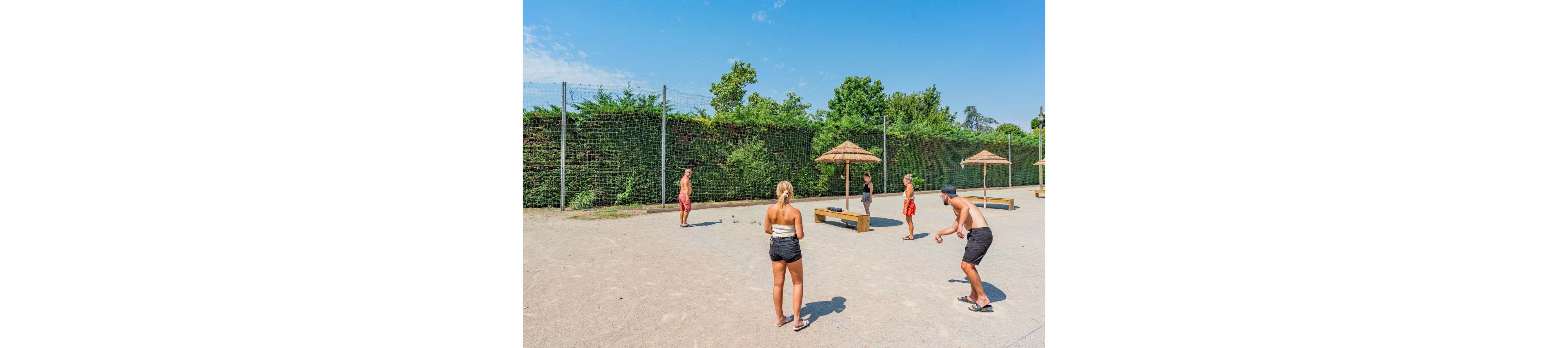 A group of young adults play a ball game on a sandy outdoor court. They stand in a loose circle under a clear blue sky, surrounded by tall green hedges and a high net fence. Wooden benches with thatched parasols provide shade in the background. The bright sunlight and casual summer clothing create a relaxed, vacation-like atmosphere.