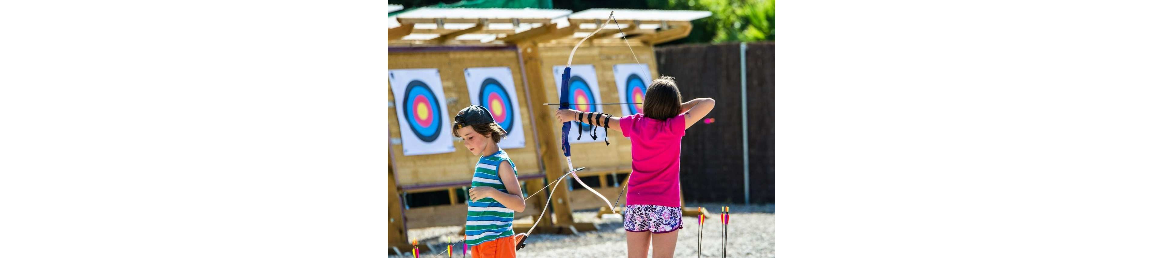 Two children are practicing archery at an outdoor range. One child stands in the foreground drawing a bow, wearing an arm guard and colorful shorts. Another child stands nearby holding a bow with several arrows on the ground. Multiple archery targets with concentric circles are set up against a wooden structure in the background. The scene is bright and sunny, suggesting a fun recreational activity or summer camp.