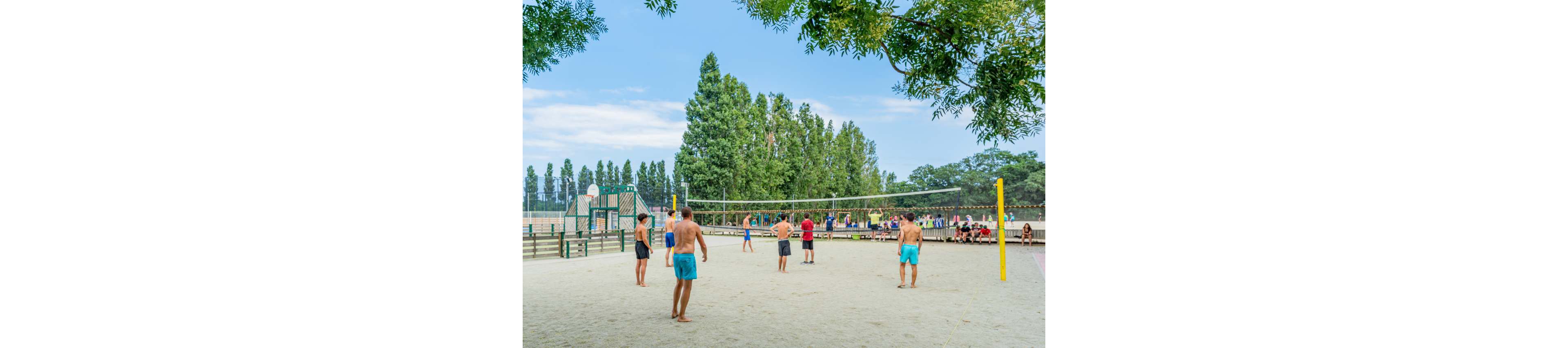A group of people play volleyball on a sand court in an outdoor park. Several players stand on either side of the net, preparing for the next play. Spectators sit on benches in the background near a fence and playground area. Tall green trees and a bright blue sky create a relaxed summer atmosphere.