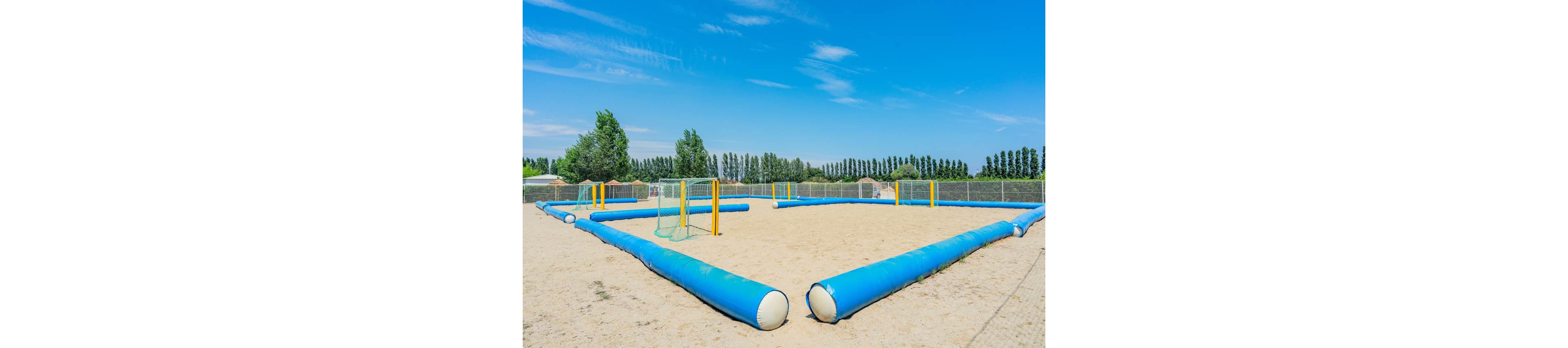 An empty inflatable sports field is set up on a sandy surface under a clear blue sky. The rectangular field is outlined with large blue inflatable tubes and includes yellow goalposts with netting. A line of tall trees and fencing surrounds the area, suggesting an outdoor recreational or training space. The bright colors and open environment create a playful, summery atmosphere.