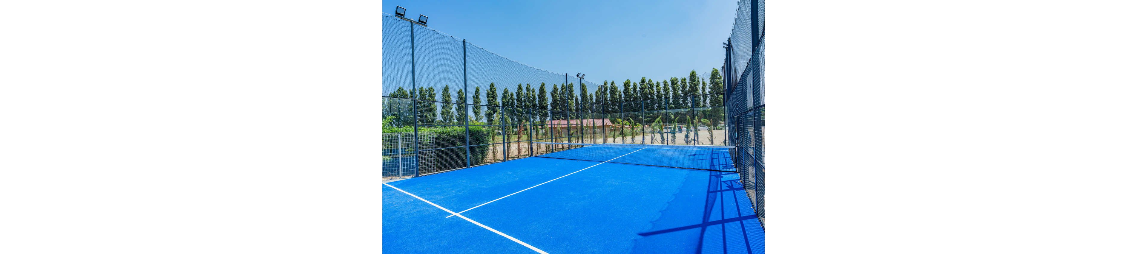An empty blue padel court is shown outdoors on a sunny day. Tall metal fences and netting surround the court, with rows of trees and greenery in the background. The bright blue playing surface contrasts with the clear sky above. The scene feels calm and ready for a game, with no players present.