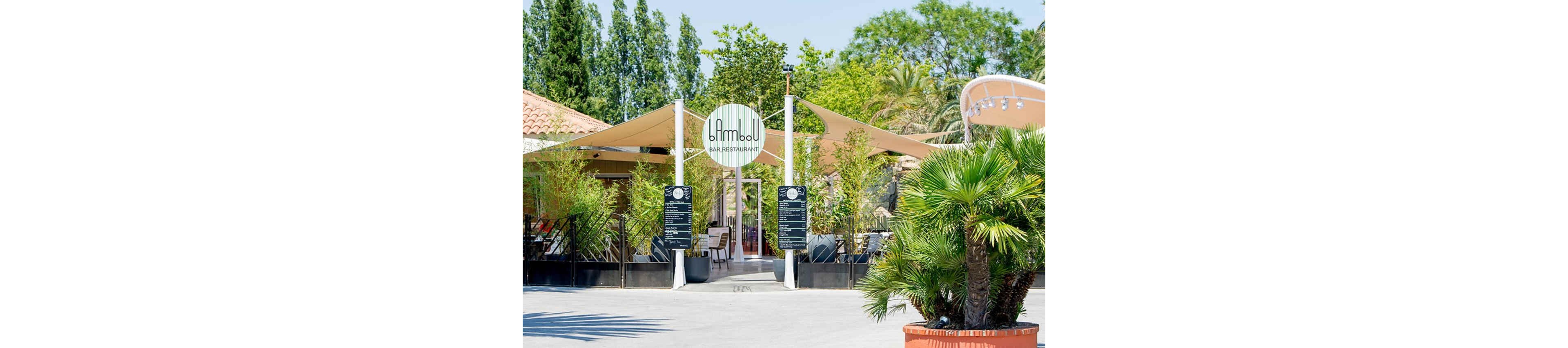 An outdoor bar restaurant with a circular sign reading "Bimbou" stands at the center of a sunny courtyard. The entrance is framed by tall bamboo plants and two black menu boards on either side. Beige shade sails stretch over the seating area, providing cover for guests. Lush greenery, including palm trees and other foliage, surrounds the venue, creating a relaxed, summery atmosphere.