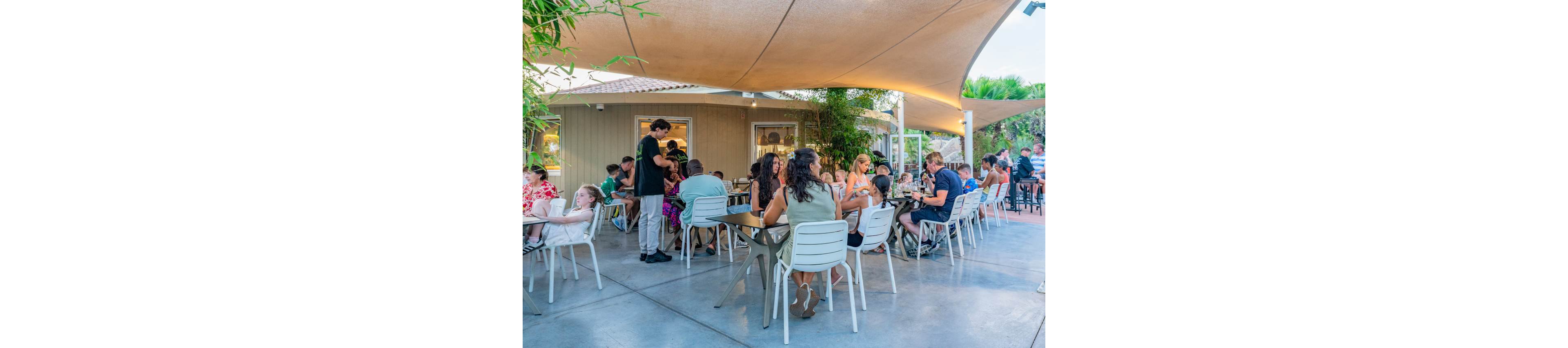 A busy outdoor cafe patio is filled with people sitting at tables and enjoying food and drinks. The seating area features white chairs and tables arranged on a smooth concrete floor under large shade canopies. Adults and children socialize in a relaxed atmosphere while a server attends to guests. Green plants and trees surround the space, and a small building with windows forms the backdrop. The scene captures a warm, casual dining experience in daylight.