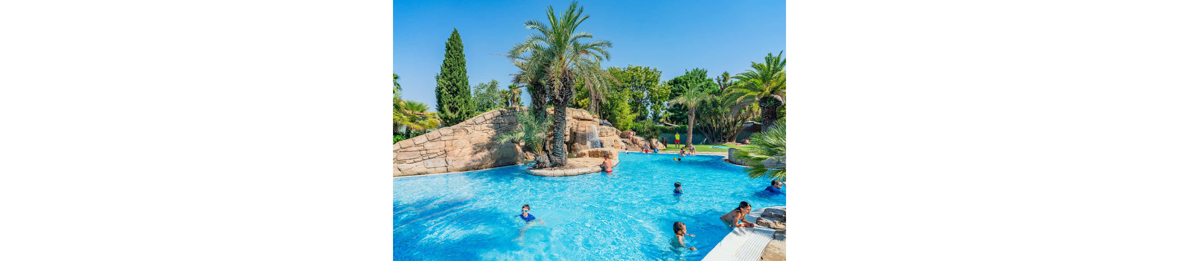 Children and adults enjoy a bright blue swimming pool in a tropical-style resort. The scene features rock formations, palm trees, and a small waterfall slide area. People are swimming, wading, and relaxing near the pool edge under a clear blue sky. The lush greenery and turquoise water create a vibrant, summery atmosphere.