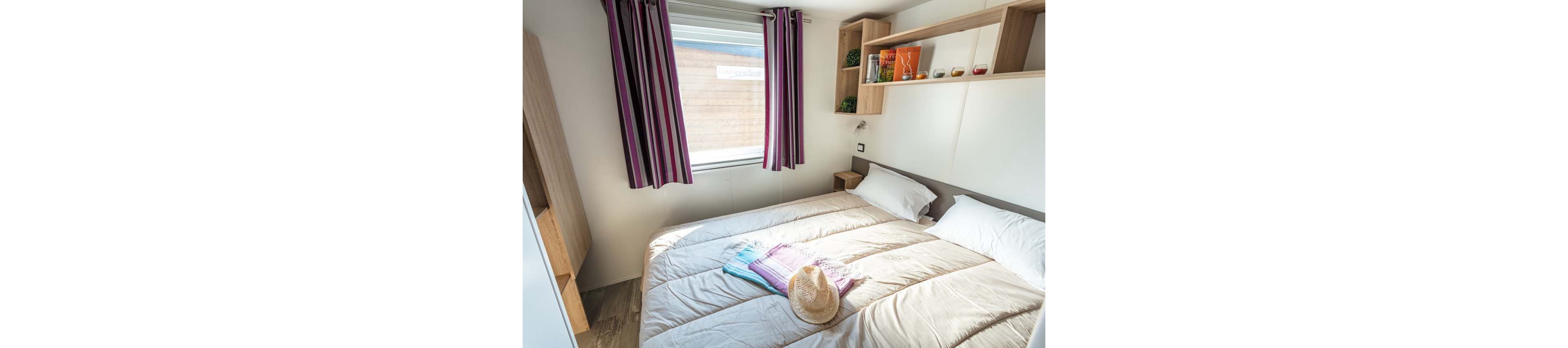 A compact bedroom features a neatly made double bed with neutral bedding. Sunlight streams through a window dressed with striped curtains, illuminating the space. A straw hat and colorful towels are placed on the bed, suggesting a vacation or beach getaway. Built-in shelves and bedside tables add storage and decorative accents, creating a tidy and inviting atmosphere.