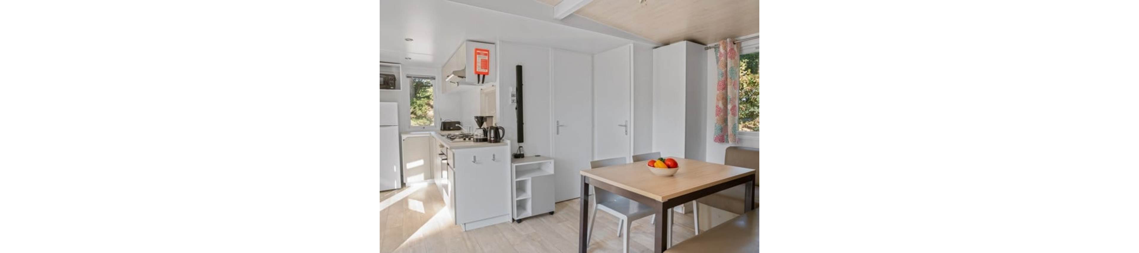 A small modern kitchen opens into a cozy dining area inside a compact home. The space features white cabinetry, a stovetop, sink, and countertop appliances along one wall. A simple wooden dining table with chairs sits in the foreground, topped with a bowl of colorful fruit. Natural light streams in through a window with patterned curtains, creating a bright and inviting atmosphere.
