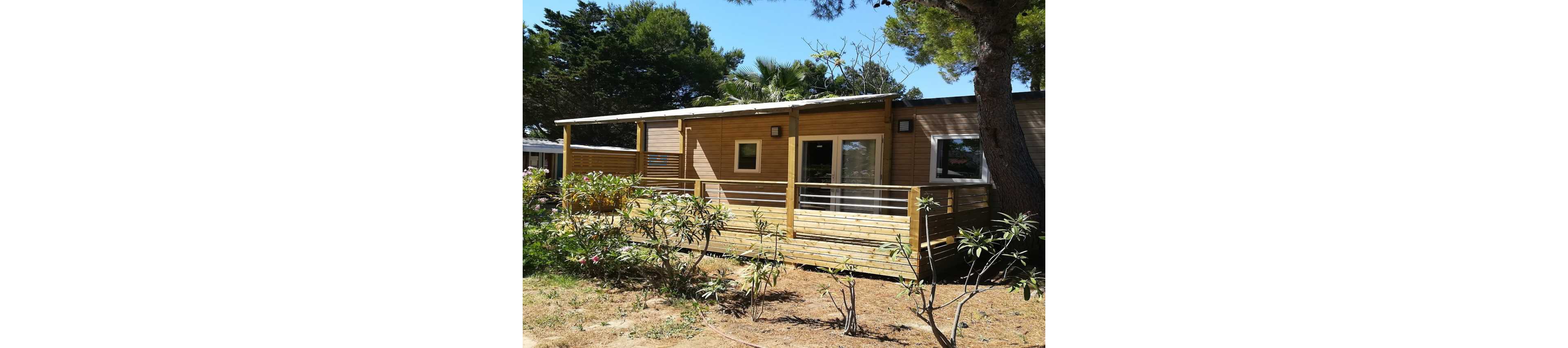 A contemporary wooden cabin with a spacious deck is set among trees and shrubs. The structure features large sliding glass doors and small square windows that open onto the sunlit terrace. A wooden railing surrounds the deck, providing a relaxed outdoor living area. Tall trees and greenery in the background create a natural, tranquil atmosphere.