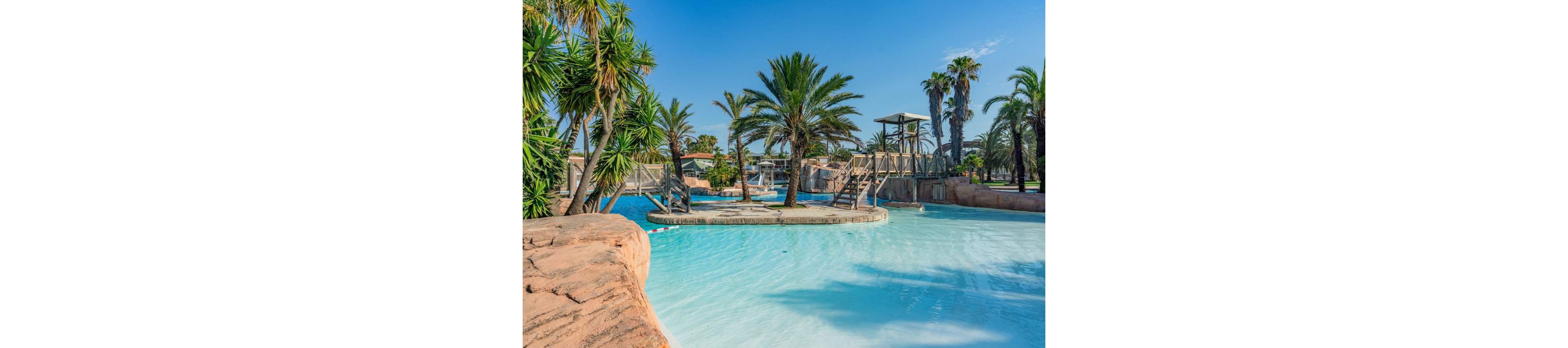 A large resort-style swimming pool is surrounded by tall palm trees and lush tropical plants. Wooden bridges and walkways cross over the bright blue water, adding structure to the leisure area. Rock-like edges frame the pool, creating a natural lagoon appearance. The scene is sunlit under a clear blue sky, suggesting a warm and relaxing vacation setting.