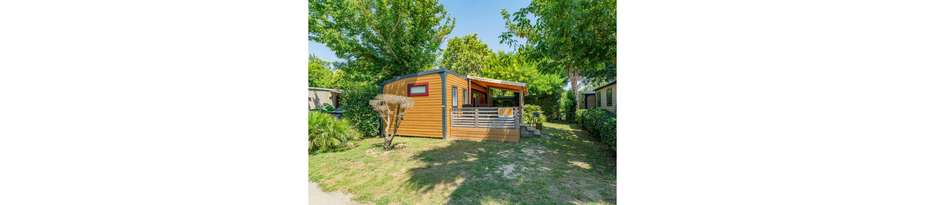 A compact wooden mobile home stands in a sunny outdoor setting surrounded by green trees and shrubs. The structure features a small covered terrace with railings and outdoor seating. Bright natural light and clear blue sky create a warm, inviting atmosphere. The scene suggests a quiet vacation spot or campsite with neighboring units visible in the background.