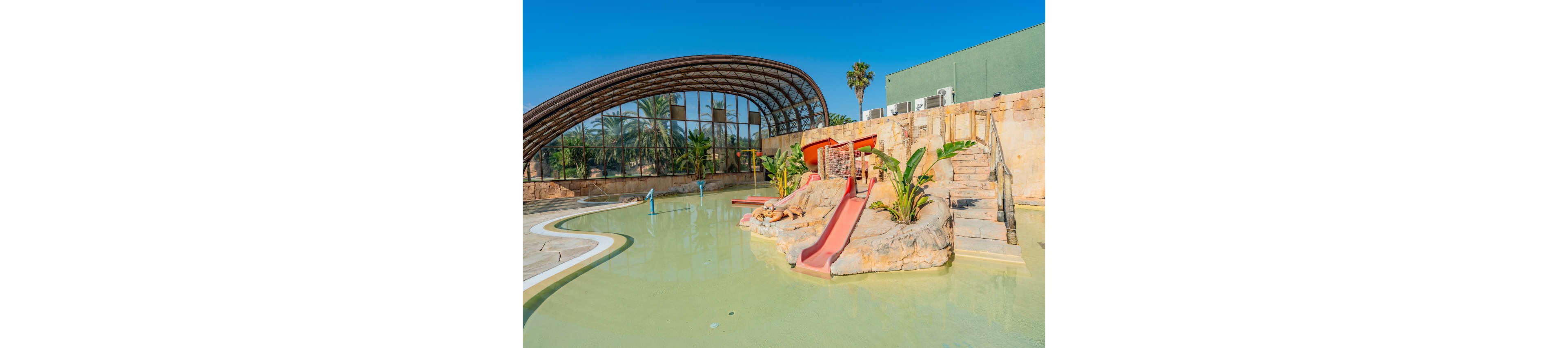 A shallow outdoor pool features small red water slides built into a rocky island. The setting appears to be a family-friendly water park with tropical plants and palm trees. In the background, a large arched glass structure encloses additional pool space, reflecting the clear blue sky. Stone walls, steps, and bright colors create a sunny, vacation-like atmosphere.