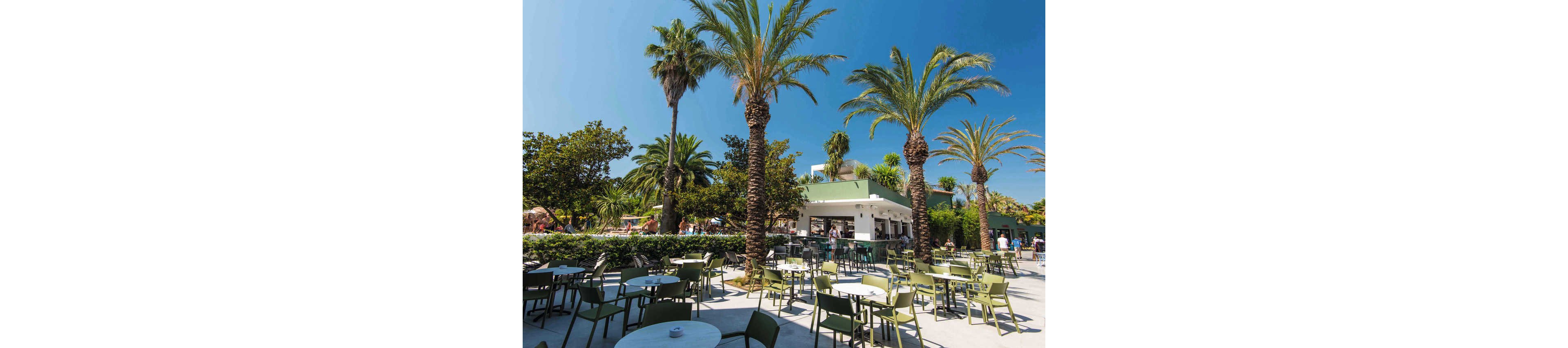 An open-air café terrace is set among tall palm trees and lush greenery. Numerous empty tables and green chairs are arranged on a sunlit patio in front of a modern white bar building. People can be seen in the background enjoying the resort facilities under a clear blue sky. The scene evokes a relaxed holiday atmosphere in a warm, tropical or Mediterranean climate.