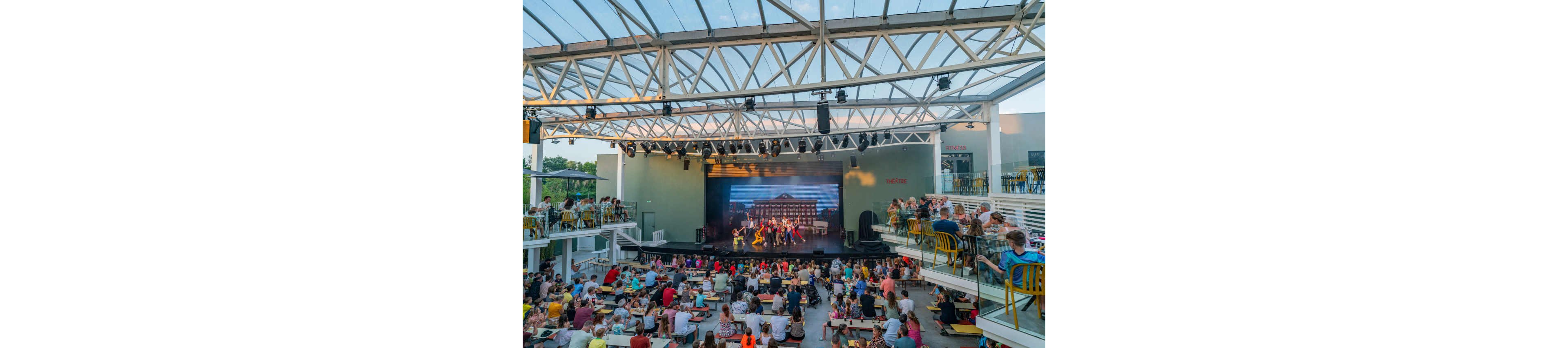 An outdoor theater hosts a live stage performance with dancers in front of a large scenic backdrop. The audience sits at tables and benches arranged in tiers under a partially covered, open-air structure. Bright daylight and clear skies create a relaxed summer atmosphere. The scene emphasizes communal entertainment, architecture, and vibrant cultural activity.