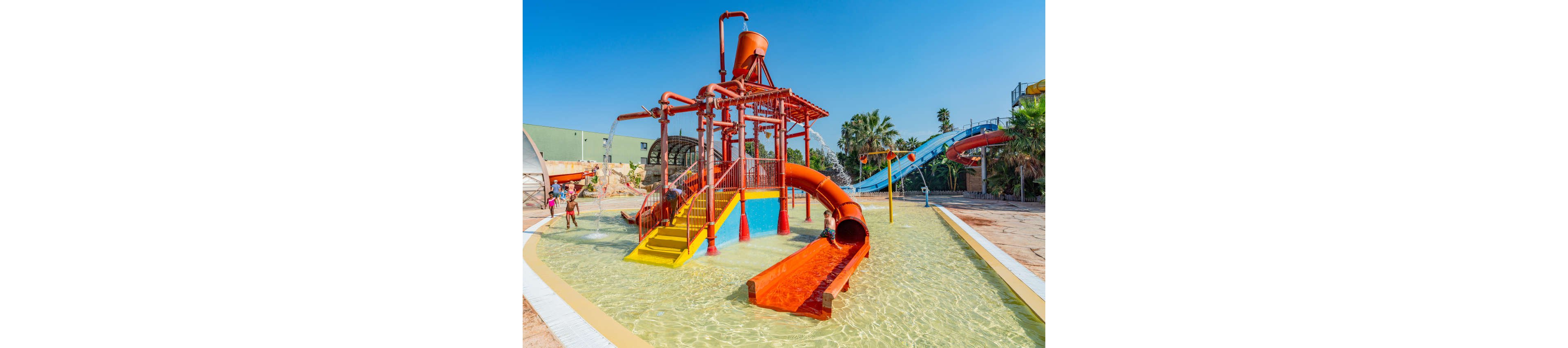 A bright outdoor water playground features multiple orange slides and climbing structures in shallow water. Children play and explore the interactive pipes and platforms under a clear blue sky. Palm trees and additional water slides are visible in the background, suggesting a larger water park. The scene feels sunny, fun, and family-friendly.