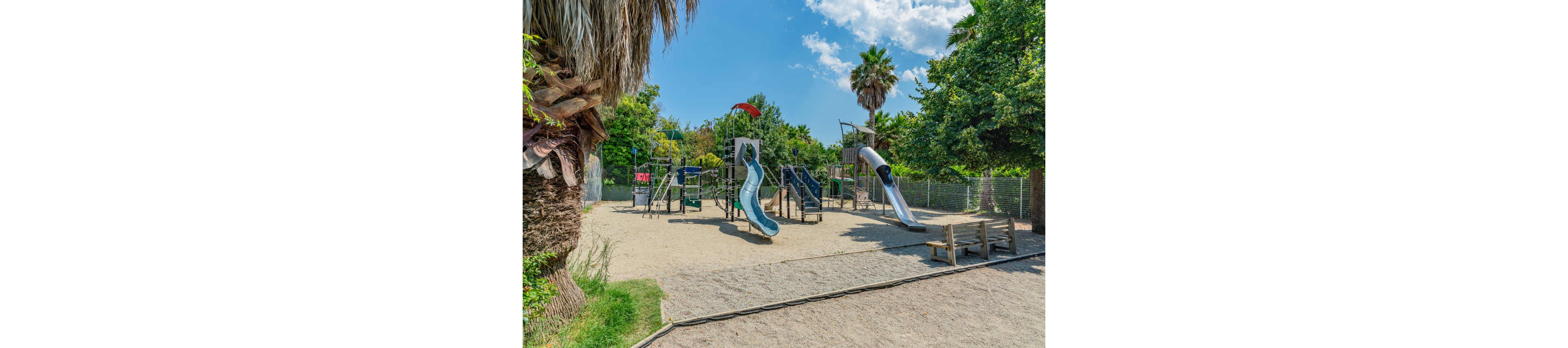 An empty outdoor playground features multiple slides and climbing structures set in a sandy play area. Tall palm trees and lush green foliage surround the fenced playground, creating a natural backdrop. The sky is bright and mostly clear, suggesting a warm, sunny day. Wooden benches sit near the play area, offering seating for parents or caregivers.