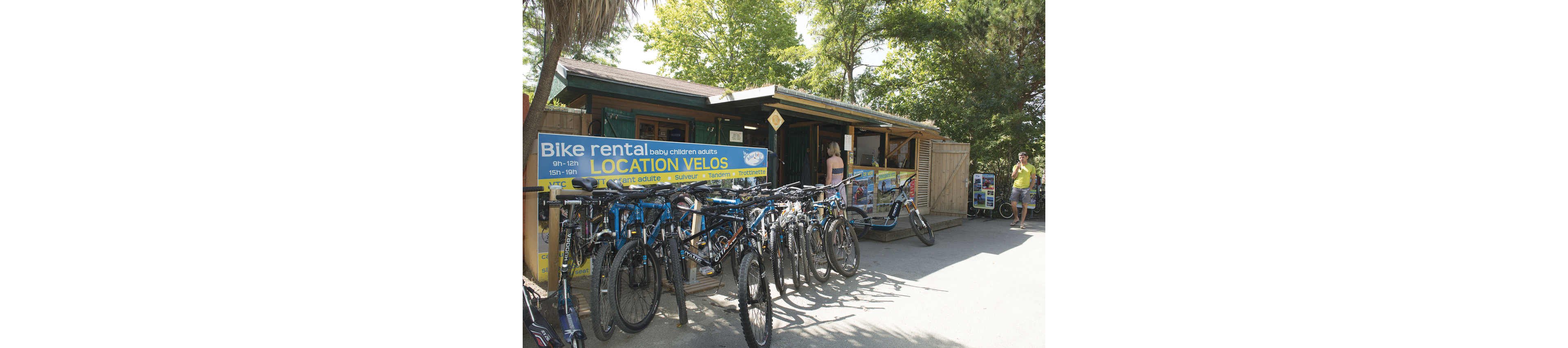 A row of bicycles is lined up in front of a small outdoor bike rental shop. The wooden kiosk is surrounded by trees and greenery, creating a relaxed holiday atmosphere. A large blue and yellow sign reads "Bike rental" and "LOCATION VELOS" with additional smaller text about services and times. Several adults stand near the kiosk, some in casual summer clothing, suggesting a warm, sunny day.