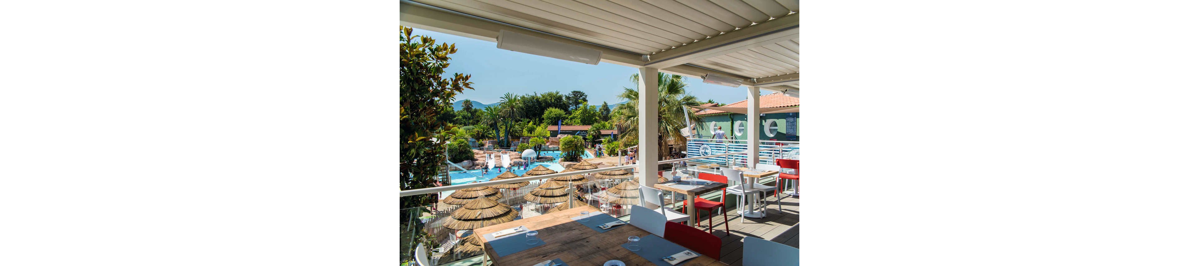 An open-air terrace restaurant overlooks a lively resort swimming pool area. Set with wooden tables, colorful chairs, and neatly arranged place settings, the dining space sits under a shaded pergola. Below, guests enjoy multiple pools, waterslides, and thatched umbrellas surrounded by palm trees and lush greenery. The bright daylight and clear blue sky create a relaxed, summery vacation atmosphere.