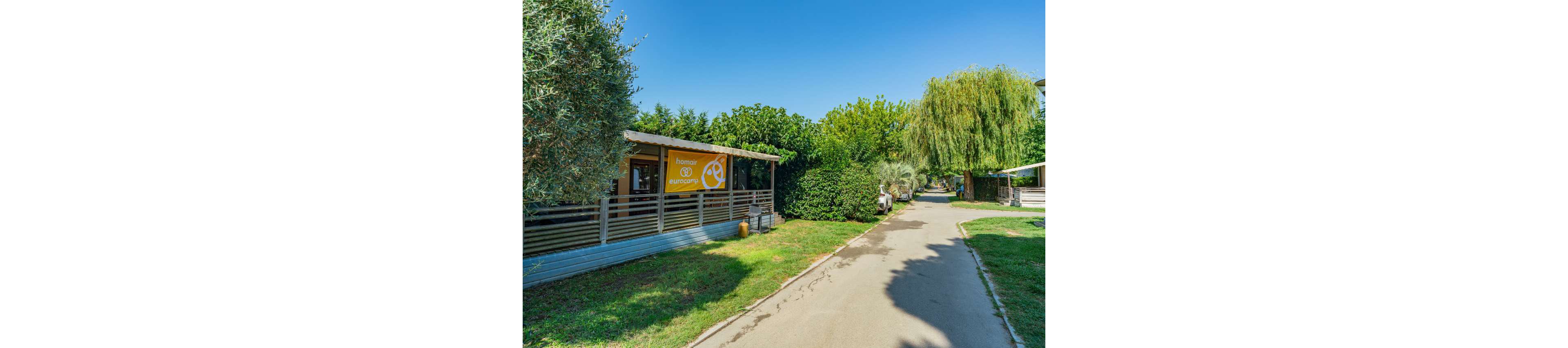 A bright summer scene shows a paved pathway running through a green campsite. On the left, a small wooden reception or service building with a yellow sign stands behind a railing. Lush trees, shrubs, and well-kept grass line both sides of the path, creating a shaded, inviting atmosphere. A person can be seen in the distance near another structure, adding a sense of scale and activity. The clear blue sky enhances the warm, holiday mood of the outdoor setting.