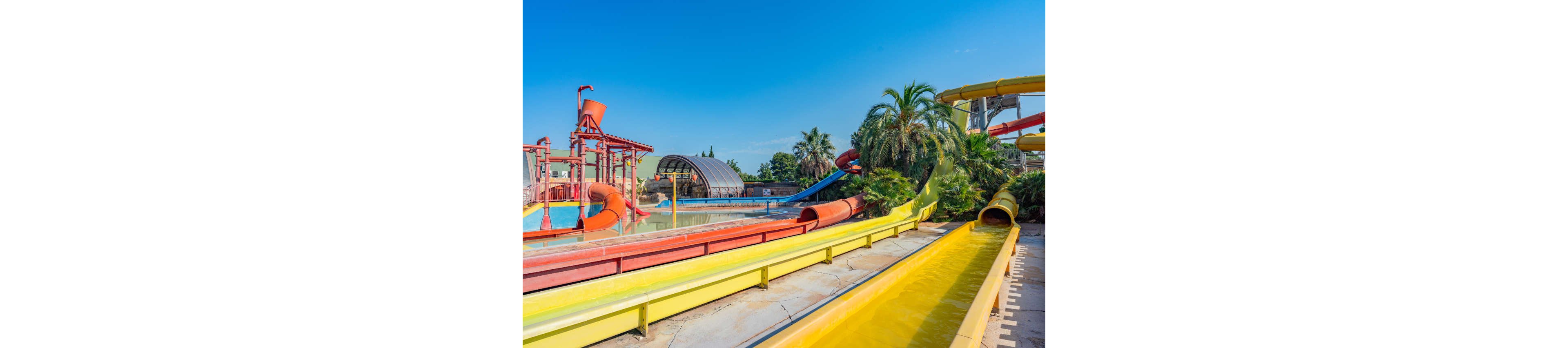 A vibrant outdoor water park features multiple colorful water slides and splash structures. The scene is set under a clear blue sky, suggesting a warm, sunny day. Palm trees and lush greenery surround the slides, adding a tropical atmosphere. The empty slides and calm water channels create a clean, inviting recreational setting.