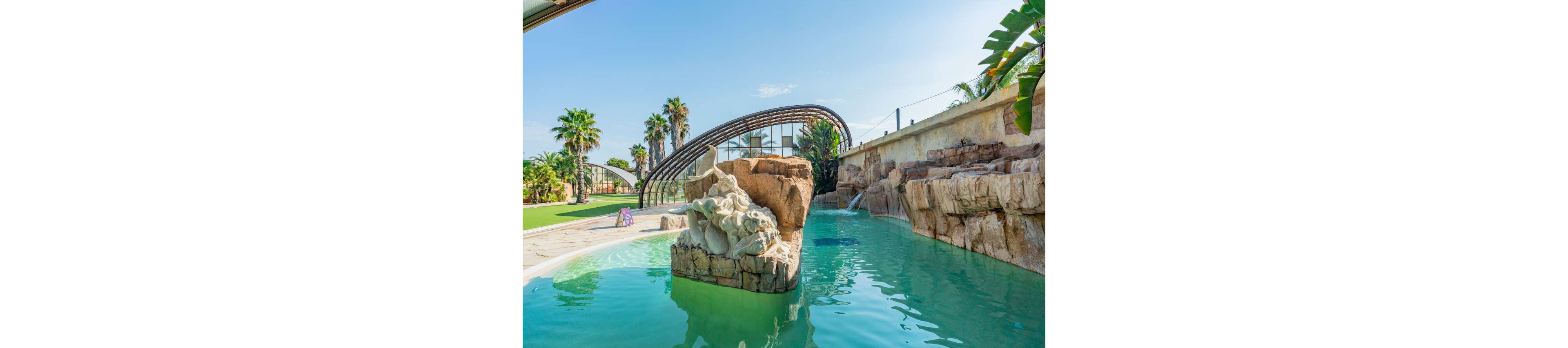 An outdoor resort-style swimming pool features a central stone sculpture rising from turquoise water. A rock wall with integrated waterfall lines one side of the pool, creating a natural grotto effect. In the background, a curved glass-and-metal structure arches over part of the pool area, framed by palm trees and manicured lawn. The bright daylight and clear blue sky give the scene a warm, tropical vacation atmosphere.