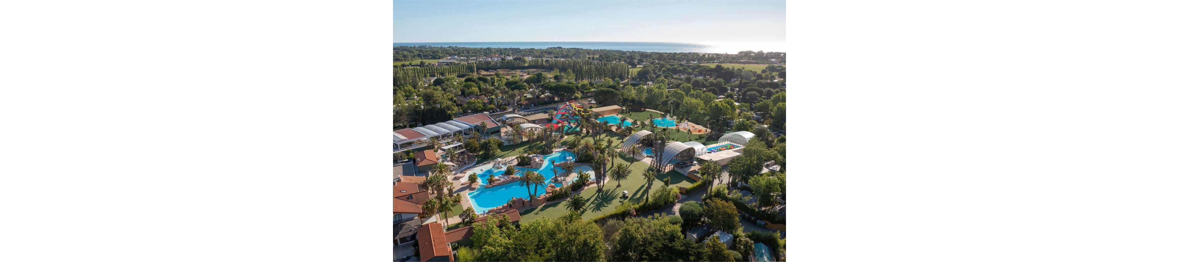 A large holiday resort with multiple swimming pools and colorful water slides is seen from an aerial perspective. The complex includes covered pool areas, open-air pools, and a central water playground surrounded by palm trees. Residential-style buildings and lush green landscaping frame the aquatic facilities. In the distance, dense tree lines and a shimmering coastline meet the horizon under clear skies.
