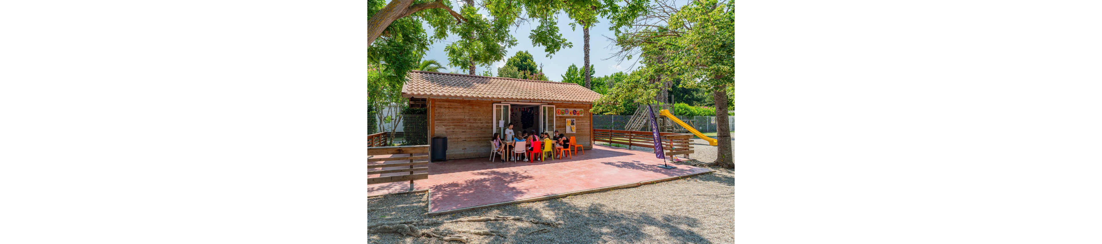 A group of children sit around tables outside a small wooden clubhouse. The scene takes place in a sunny outdoor recreation area with trees providing shade. A playground with a slide and climbing structure is visible in the background behind a fence. Brightly colored chairs and a poster on the cabin wall add to the lively, summery atmosphere.