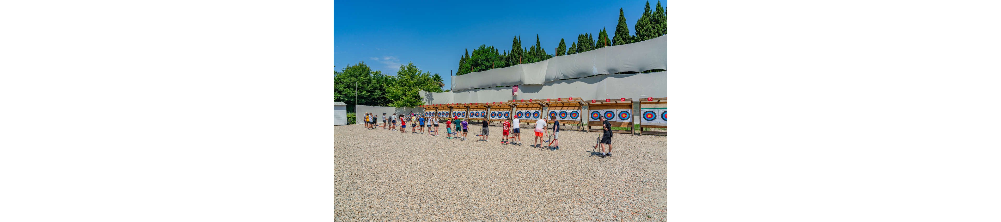 A large group of people practice archery at an outdoor shooting range. Participants stand in a line on a gravel surface, each aiming toward colorful circular targets mounted on a long wooden structure. The scene is set under a clear blue sky with trees and tall barriers surrounding the range. The image conveys a recreational, instructional atmosphere with a focus on group sports and skill training.