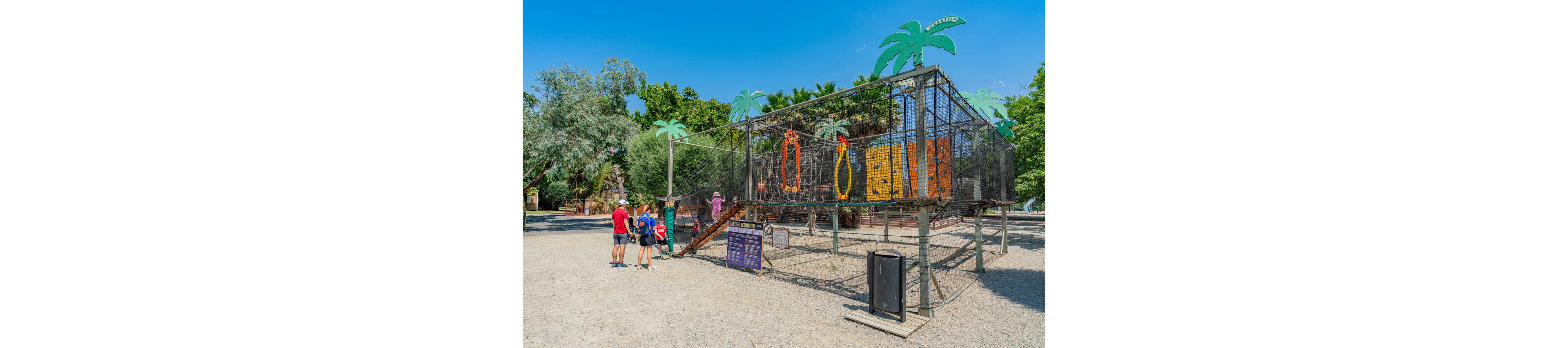 A family stands in front of a large outdoor cage-like exhibit decorated with bright animal silhouettes. The structure is topped with playful palm tree cutouts and surrounded by gravel ground in a park setting. Several adults and children observe the installation while one child climbs a small ramp to the entrance. Lush green trees and a clear blue sky create a sunny, relaxed atmosphere around the attraction.