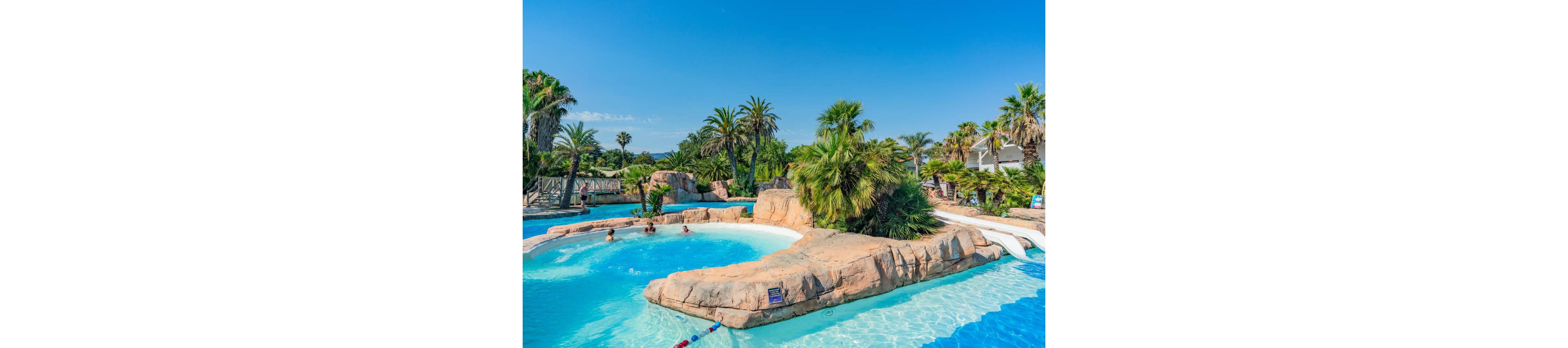 A large tropical-style swimming pool features curving blue water around artificial rock formations. Palm trees and lush greenery surround the pool, creating a vacation resort atmosphere under a clear blue sky. Several people relax and swim in the water near the rocks and shallow areas. White water slides and separated swimming zones add to the fun, family-friendly setting.