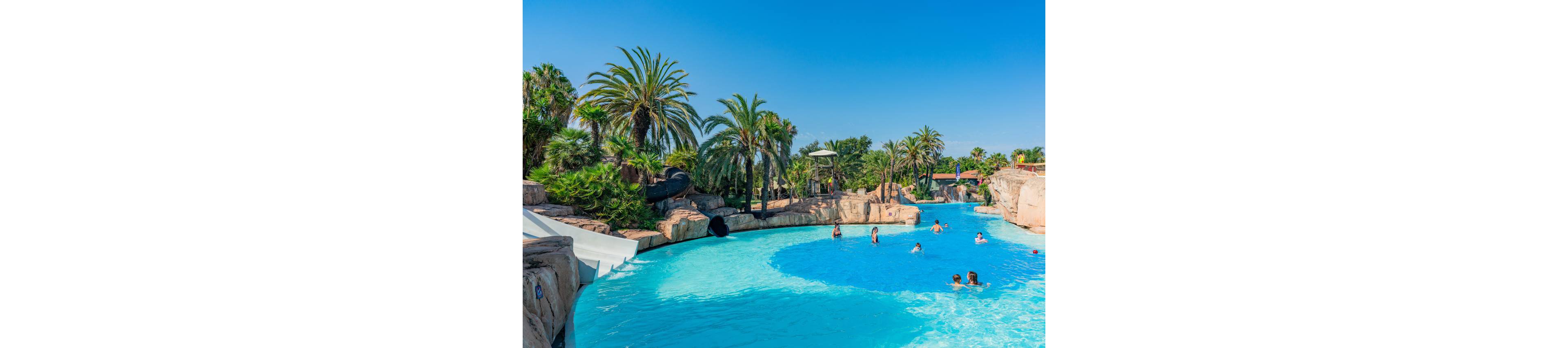 A large outdoor swimming pool curves through a tropical-themed water park. Children and adults play in the bright blue water near rock formations and water slides. Palm trees and lush greenery surround the pool under a clear blue sky. The scene suggests a warm, sunny vacation atmosphere with relaxed family fun.