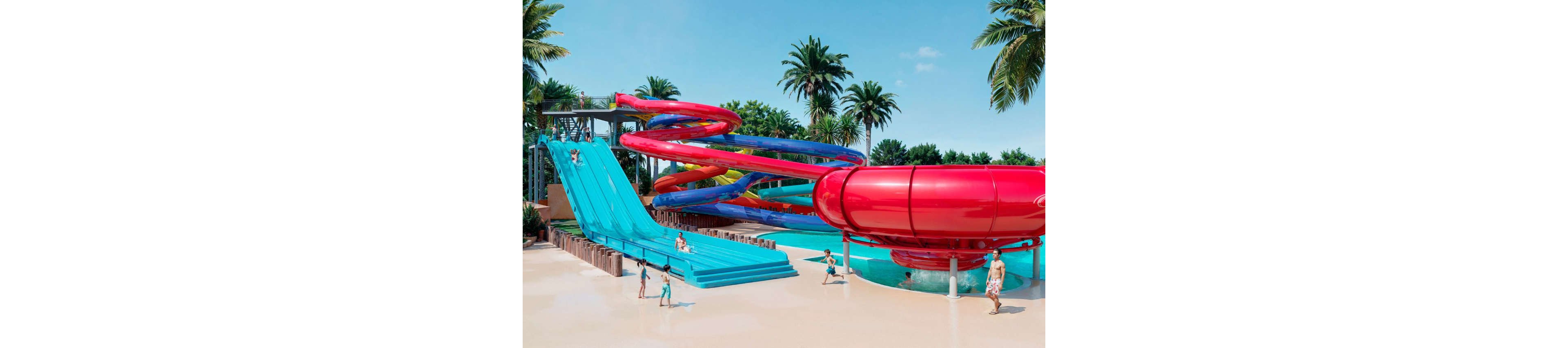 A vibrant water park features large blue and red slides surrounded by palm trees. Children and adults enjoy the attractions, with some climbing the stairs and others running across the shallow water area. The twisting enclosed slide and wide multi-lane slide dominate the scene under a clear blue sky. The overall atmosphere is playful, energetic, and summery.
