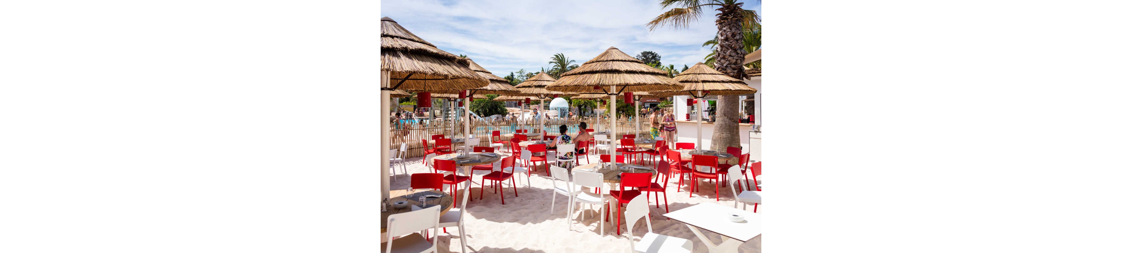 An open-air café with red and white chairs is set directly on white sand under thatched umbrellas. People sit at tables enjoying drinks and conversation while others walk near the bar area. In the background, a swimming pool and palm trees create a relaxed resort atmosphere. Bright daylight and clear skies give the scene a summery, vacation mood.