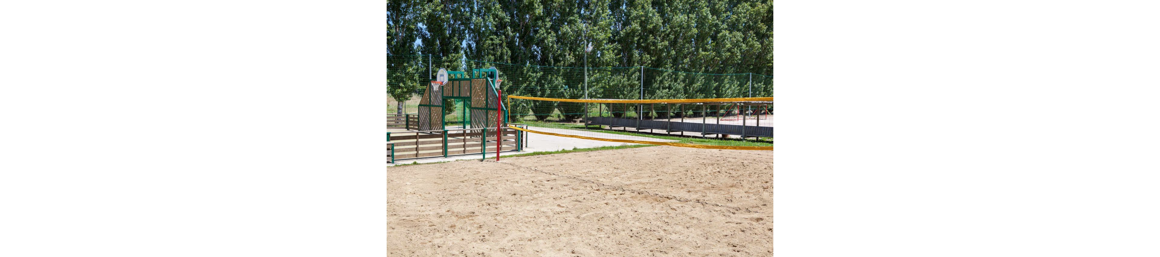 A group of children play volleyball on a sandy outdoor court. Two kids jump near the net to hit the colorful ball while others watch and wait to participate. The court is surrounded by a tall green fence, with trees and a playground basketball hoop visible in the background. Bright summer clothing and clear skies create a lively, energetic atmosphere.