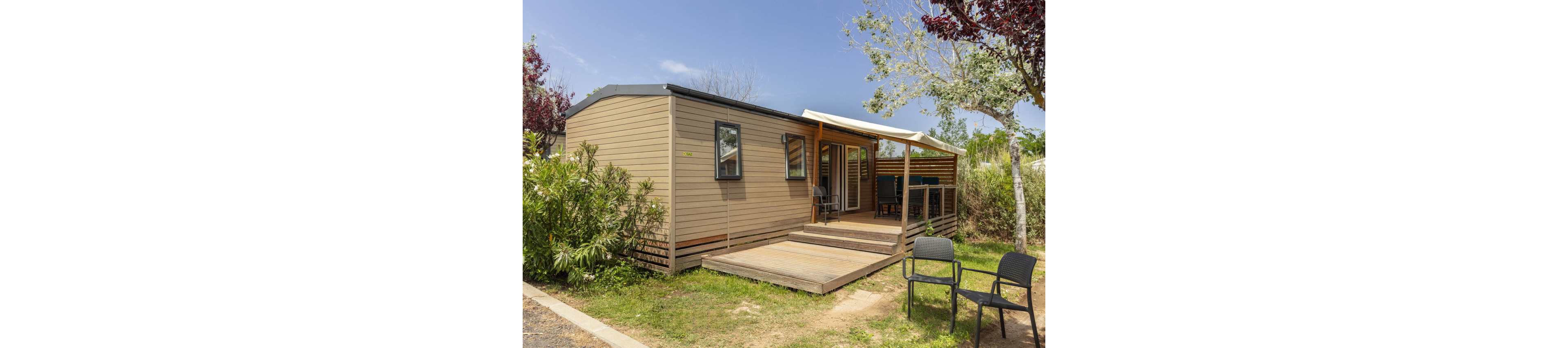 A contemporary mobile home sits on a small plot surrounded by greenery and trees. The structure features light wood siding, a flat dark roof, and a raised wooden deck at the entrance. A covered terrace area holds an outdoor dining table and chairs, while two additional chairs are placed on the grass nearby. The scene is lit by bright daylight under a clear blue sky, suggesting a relaxed holiday or campsite setting.