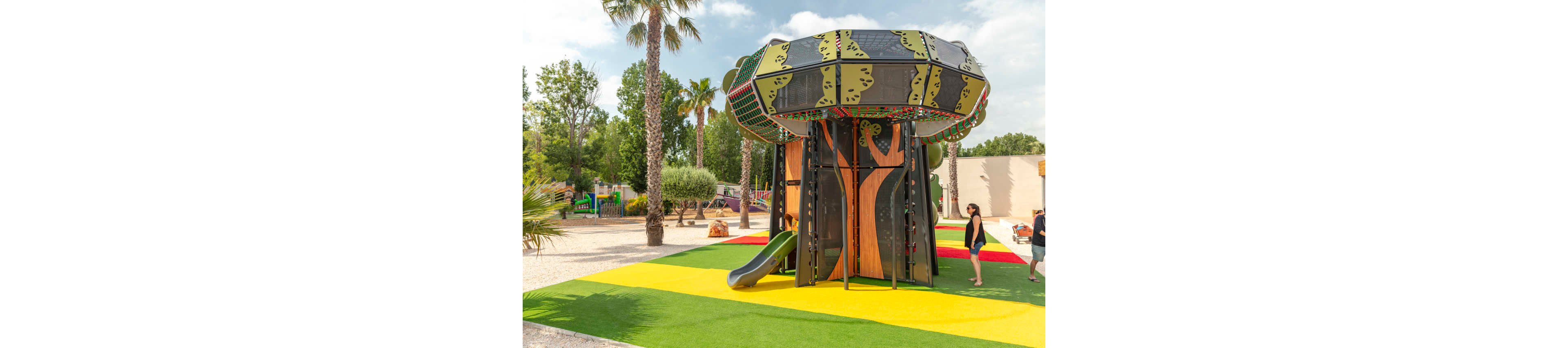 A large tree-themed playground structure with slides stands on a brightly colored rubber surface. The play area features bold green, yellow, and red stripes beneath the equipment. Palm trees and other greenery surround the playground, creating a sunny outdoor park setting. A few adults and children walk nearby, enjoying the recreational space.