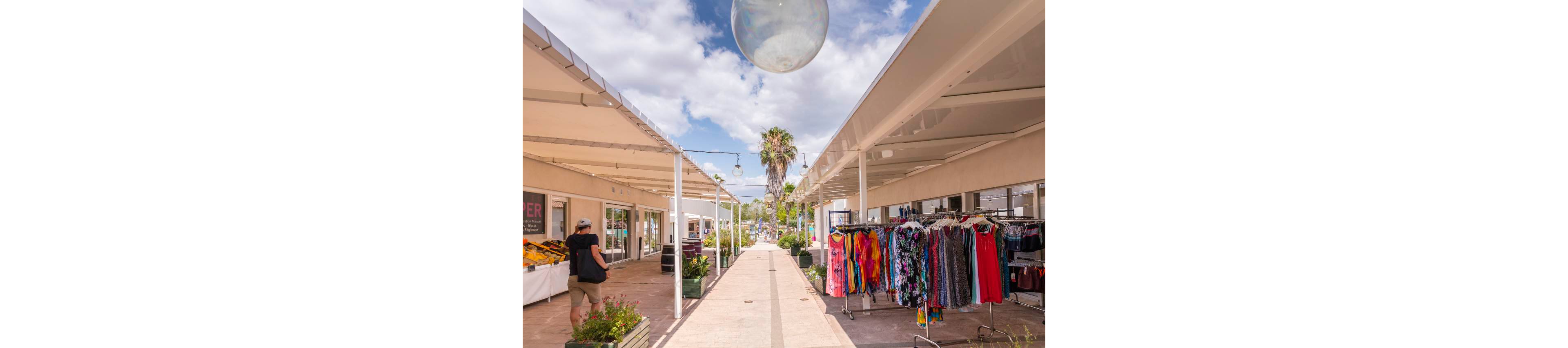 An open-air shopping arcade features racks of colorful dresses and garments on the right side of the walkway. A person with a hat and backpack browses near a produce or goods stall on the left. White canopy structures line both sides of the path, leading toward a palm tree and bright sky with scattered clouds. The scene suggests a relaxed, casual shopping experience in warm weather.