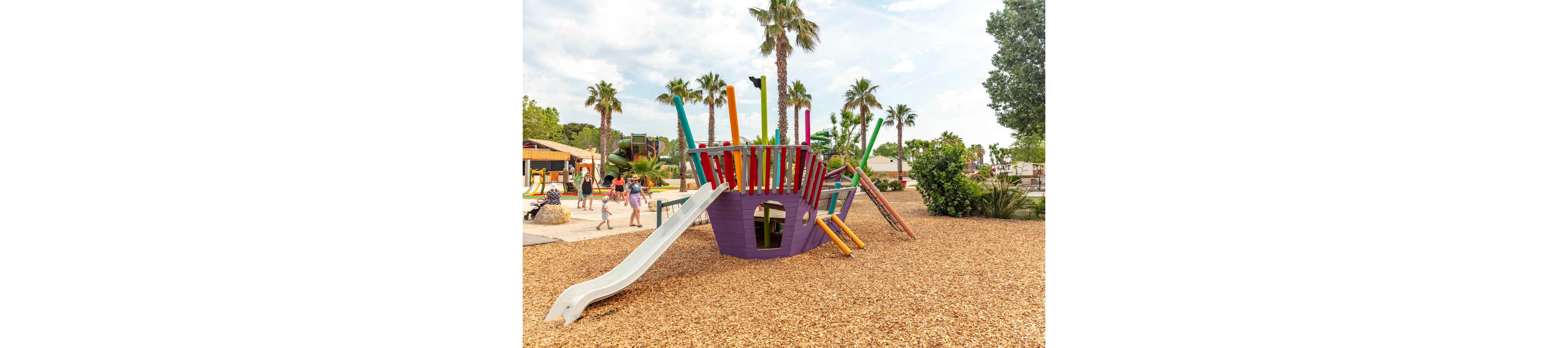 A brightly colored playground structure shaped like a ship stands on a bed of wood chips. The play area features a white slide, purple and red panels, and tall multicolored poles. Children and adults walk nearby, enjoying the outdoor recreation space. Palm trees and greenery surround the playground under a partly cloudy sky, creating a warm, family-friendly atmosphere.