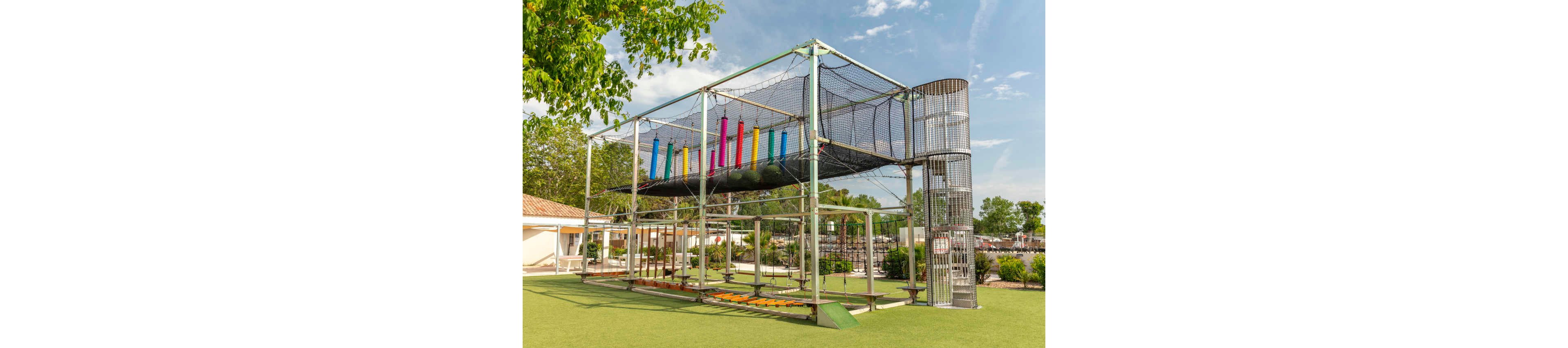 A large metal adventure playground structure stands on a bright green artificial grass surface. The play equipment features elevated walkways, nets, and colorful hanging cylinders for climbing and balancing. A tall cylindrical cage-like tower connects to upper platforms and safety nets. Trees, low buildings, and a clear blue sky with scattered clouds create a sunny, inviting outdoor setting.
