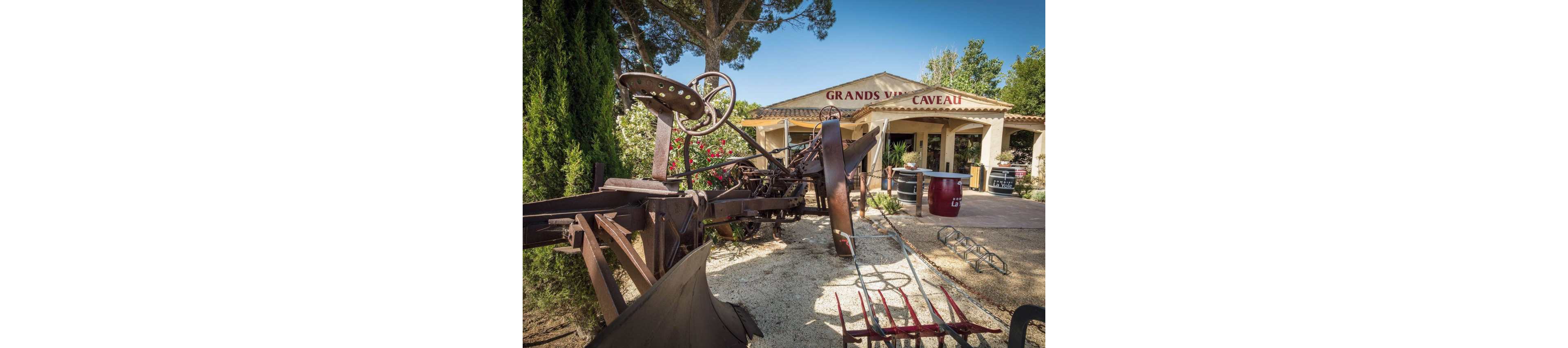 A rustic winery courtyard features large pieces of vintage agricultural machinery in the foreground. Behind the equipment stands a light-colored building with a tiled roof and the words "GRANDS VINS CAVEAU" on the facade. Tall trees and green shrubs frame the scene, adding a natural backdrop. Wine barrels and outdoor furniture near the entrance suggest a welcoming tasting area for visitors.