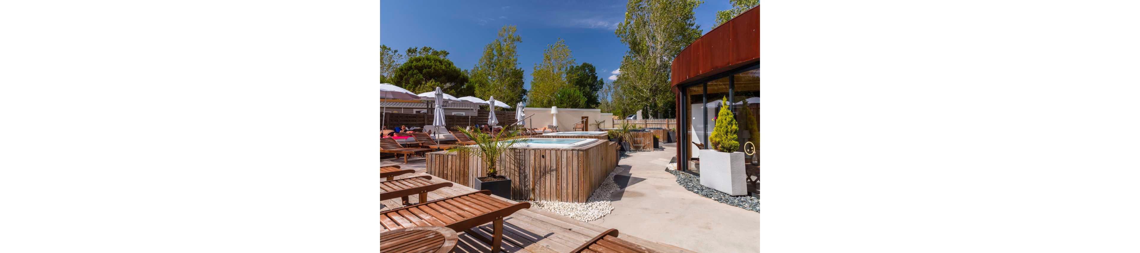 An outdoor spa area features a raised rectangular hot tub surrounded by wooden cladding. Numerous wooden sun loungers are arranged on a spacious deck under a clear blue sky. White umbrellas shade a seating area where people relax in the background. Modern landscaping with potted plants and a curved building facade adds to the resort-like atmosphere.