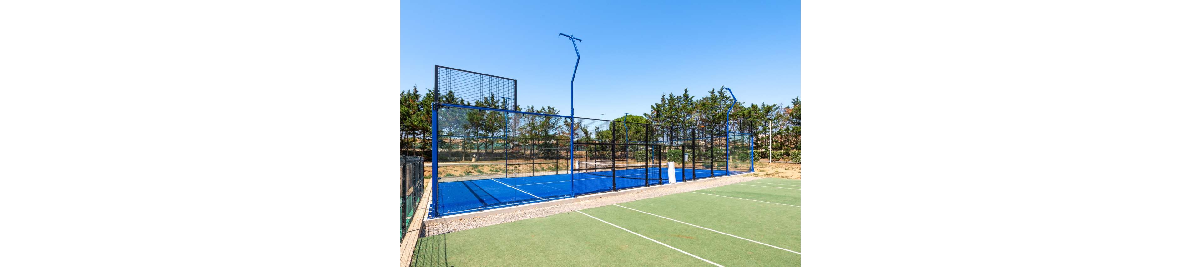 Image of an outdoor sports facility featuring a bright blue padel court enclosed by high metal fencing. The padel court stands next to a green tennis court with white boundary lines. Trees and shrubs form a natural backdrop under a clear blue sky. The scene is empty of players, emphasizing the clean, well-maintained surfaces and open recreational setting.