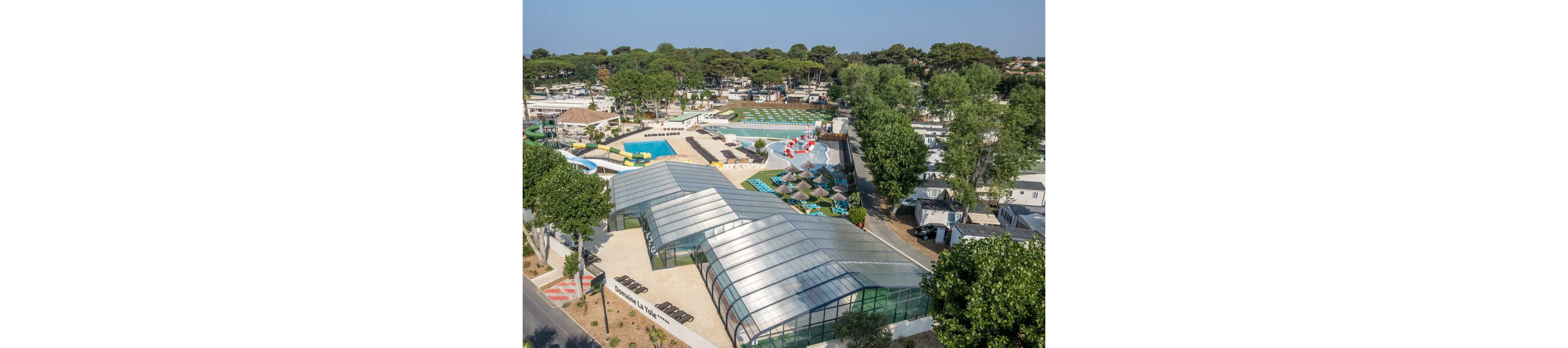 A large holiday campsite with an extensive waterpark is seen from above on a sunny day. The foreground features a modern glass-roofed building next to a tree-lined road. Behind it, multiple outdoor swimming pools, water slides, and sun loungers are arranged in neat sections. Rows of mobile homes and cabins extend into the background, surrounded by tall green trees. The overall scene suggests a busy family vacation resort with many recreational facilities.